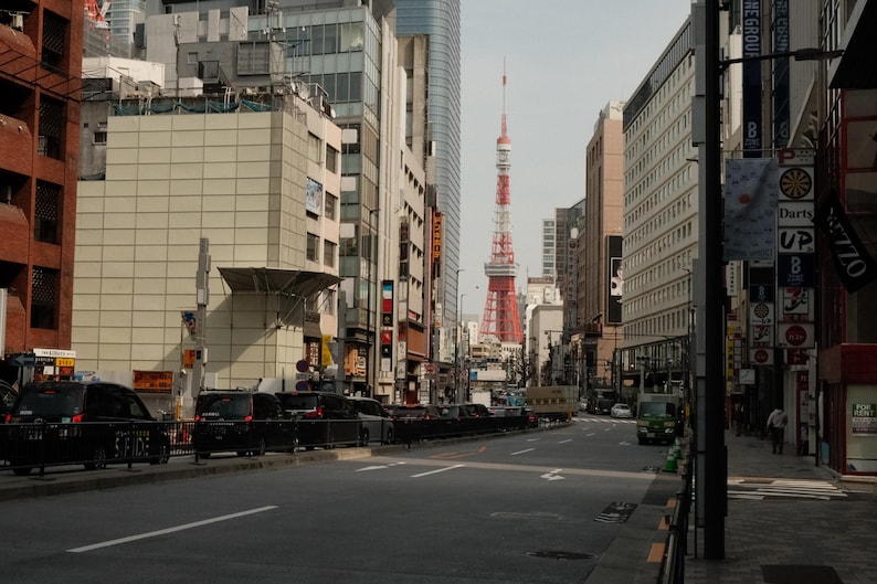 Puede incluir: Una vista de una calle de la ciudad de Tokio, Jap&oacute;n, con una alta torre roja y blanca en la distancia. La torre es visible entre los edificios y es el punto focal de la imagen. La calle est&aacute; bordeada de edificios y coches.