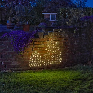 May include: Two illuminated Christmas tree shapes made of white lights against a brick wall at night.