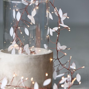 May include: A decorative LED light garland with white leaf-shaped accents and warm white lights. The garland is wrapped around a clear glass cylinder and a concrete base, creating a soft, ambient glow. Ideal for home decor.