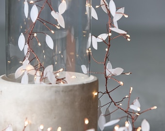 May include: A decorative LED light garland with white leaf-shaped accents and warm white lights. The garland is wrapped around a clear glass cylinder and a concrete base, creating a soft, ambient glow. Ideal for home decor.