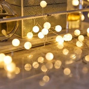 May include: A string of small, round, glowing white lights, resembling fairy lights, are draped across a surface. The lights are the focal point, casting a warm, soft glow. Other objects, including a decorative box, are visible in the background.