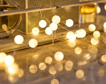May include: A string of small, round, glowing white lights, resembling fairy lights, are draped across a surface. The lights are the focal point, casting a warm, soft glow. Other objects, including a decorative box, are visible in the background.