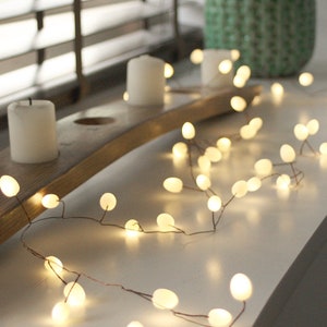 May include: A string of warm white fairy lights with round bulbs, draped over a wooden base with three white candles. The lights are strung on a copper wire and are ideal for adding a touch of ambience to any room.