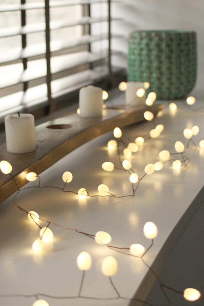 May include: A string of warm white fairy lights with round bulbs, draped over a wooden base with three white candles. The lights are strung on a copper wire and are ideal for adding a touch of ambience to any room.