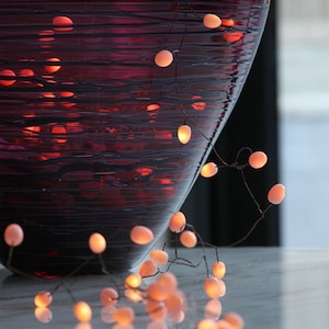May include: A purple glass vase with a string of warm white lights wrapped around it. The lights are strung on a thin wire and are spaced evenly apart. The vase is sitting on a white surface.