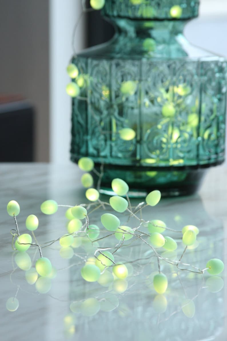 May include: A string of fairy lights with green frosted bulbs. The lights are strung on a silver wire and are wrapped around a decorative green glass vase.