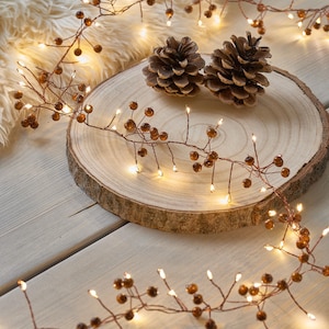 May include: A decorative garland with warm white LED lights and brown berry-like accents. The garland is draped over a wooden slice with two pine cones. The lights are on, creating a cozy, festive atmosphere.