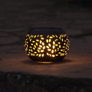 May include: A black, spherical lantern with a pattern of leaf-shaped cutouts. The interior glows with a warm yellow light, creating a soft, inviting ambiance. The lantern is made of a dark material and sits on a stone surface.
