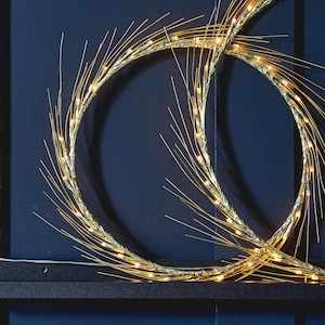 May include: Gold-colored, circular decorative lights with small, warm white LED bulbs. The lights are arranged in a wreath-like shape with thin, spiky accents. The background is a dark blue.
