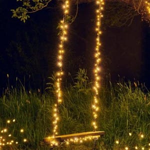 May include: A wooden swing seat suspended by two thick black ropes, both wrapped in long strands of warm white fairy lights. The lights extend upward into the branches of a tree and illuminate the seat and the surrounding dark grassy area.