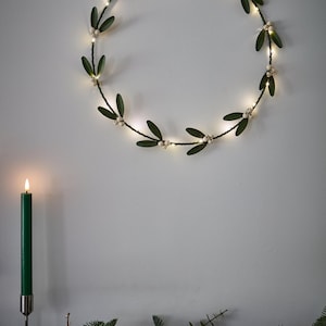 May include: A lighted green wreath with white berries hangs on a white wall. A green candle in a silver holder is lit on the left. The wreath is decorated with small white lights, creating a festive holiday decoration.