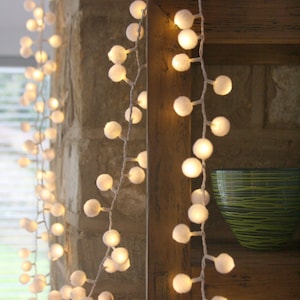 May include: White pom pom string lights with warm white LED bulbs, draped over a wooden shelf and a brick wall.