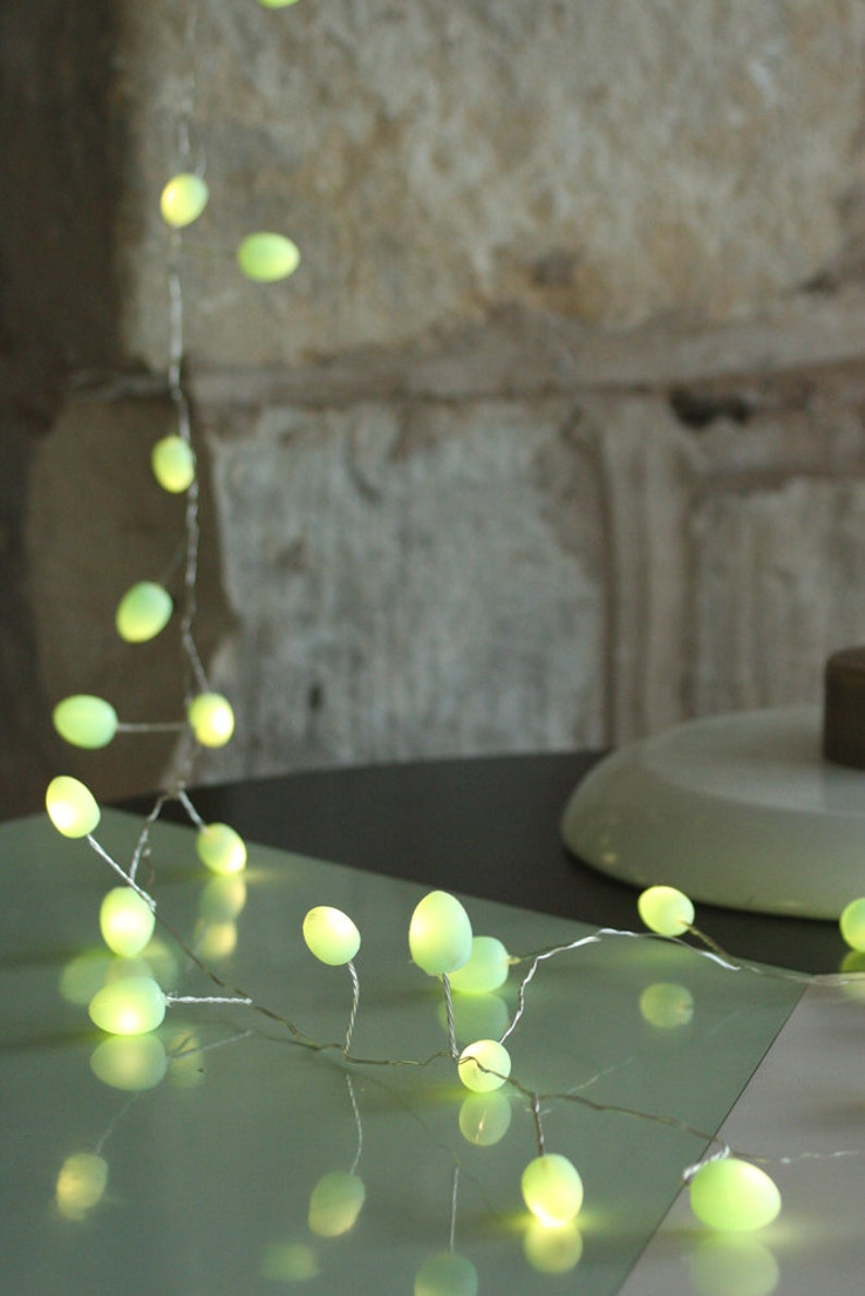 May include: A string of fairy lights with green, oval-shaped bulbs. The lights are strung on a silver wire and are lit, casting a warm glow.