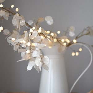 May include: A white ceramic jug with a delicate branch of white paper flowers and small warm white lights. The jug is sitting on a woven brown tray.