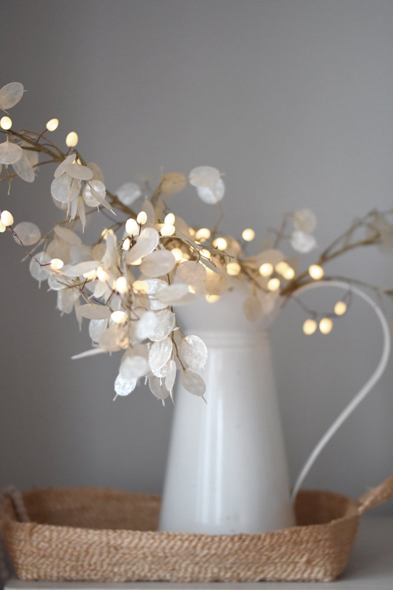 May include: A white ceramic jug with a delicate branch of white paper flowers and small warm white lights. The jug is sitting on a woven brown tray.