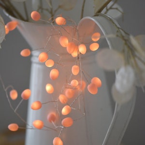 May include: A white ceramic jug with a string of warm white fairy lights wrapped around it. The lights are shaped like small, round bulbs and emit a soft glow. The jug is decorated with a sprig of dried flowers.
