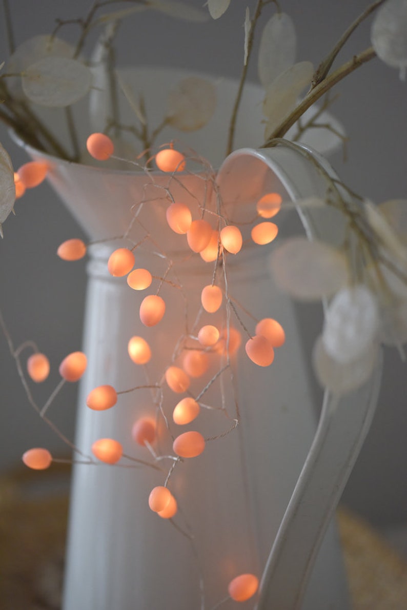 May include: A white ceramic jug with a string of warm white fairy lights wrapped around it. The lights are shaped like small, round bulbs and emit a soft glow. The jug is decorated with a sprig of dried flowers.