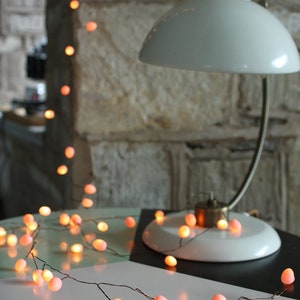 May include: A string of warm white fairy lights with pink bulbs wrapped around a table. The lights are on and glowing. A white lamp with a white shade is in the background.