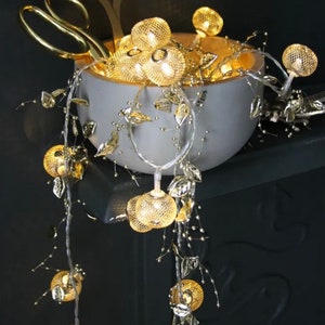 May include: A string of fairy lights with gold wire and gold mesh globe lights. The lights are draped over a white bowl with gold leaves and a pair of gold scissors.