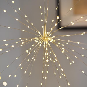 May include: A decorative starburst light fixture with numerous tiny, warm white LED lights radiating from a central point. The lights are attached to thin, flexible wires, creating a dynamic, illuminated display. The fixture is designed for indoor use.