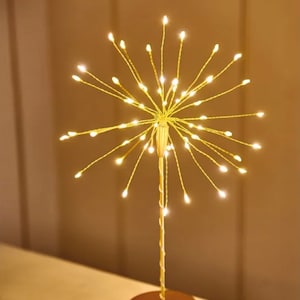 May include: A decorative gold-colored light fixture with a round base and a central stem that branches out into multiple thin wires, each tipped with a small, warm white LED light. The fixture resembles a burst of light or fireworks.