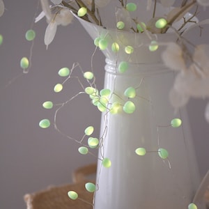 May include: A white ceramic jug with a green fairy light garland draped around it. The lights are shaped like small, round bulbs and are strung on a thin wire. The jug is sitting on a woven basket.