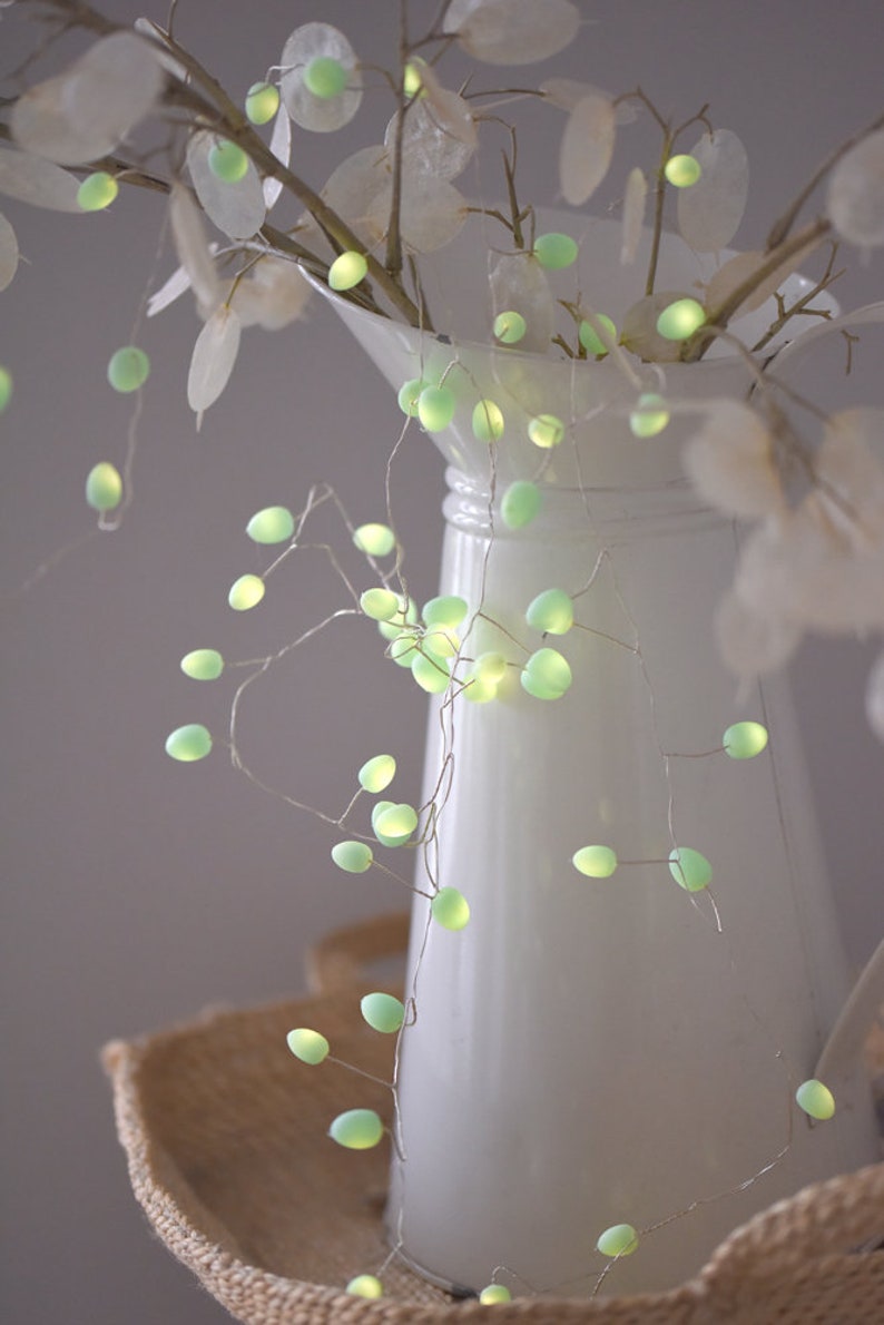 May include: A white ceramic jug with a green fairy light garland draped around it. The lights are shaped like small, round bulbs and are strung on a thin wire. The jug is sitting on a woven basket.