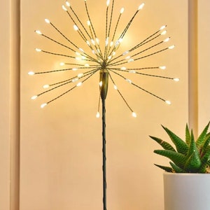 May include: A black metal tabletop light fixture with a round base and a long stem. The stem has a cluster of small white LED lights that resemble a firework display.