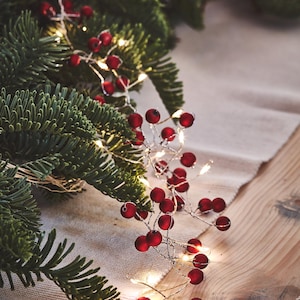 May include: A festive Christmas garland featuring faux red berries and small, warm white LED lights. The garland is intertwined with green pine branches, creating a seasonal decorative element. The lights are on, illuminating the berries.