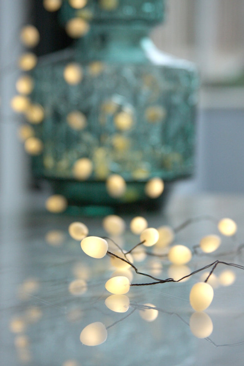 May include: A string of white fairy lights with a warm glow, draped over a textured teal vase. The lights are arranged in a random pattern, creating a whimsical and festive atmosphere.