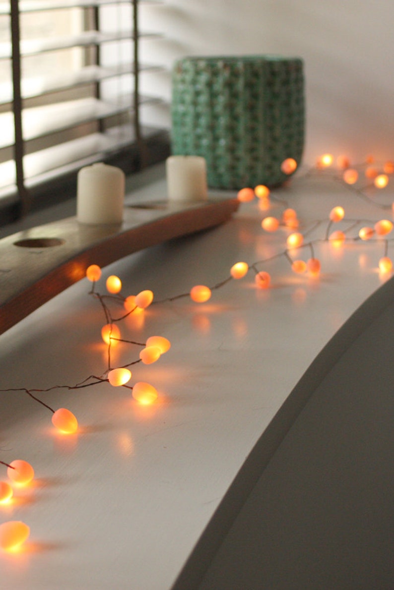May include: A string of warm white fairy lights with small, round, peach-coloured bulbs. The lights are strung on a thin wire and are draped over a white surface.