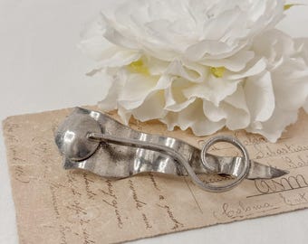 May include: A vintage silver-toned hairpin with a unique, flowing design. The hairpin features a circular end, a wavy central section, and a pointed tip. It rests on a vintage postcard with faded text, with a white flower in the background.