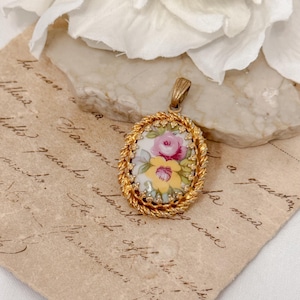 以下が含まれることがあります： ピンクと黄色のバラの花柄が描かれた白い磁器製の楕円形のカメオがついた、金色のペンダント。カメオは金色のロープ状の縁取りで囲まれています。