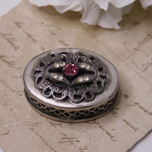 Puede incluir: Una caja de alhajas ovalada de plata con un diseño filigranado intrincado y un strass rosa en el centro.