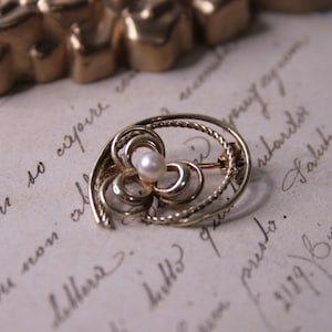 以下が含まれることがあります： 金色のヴィンテージブローチ。花のモチーフと中央のパールが特徴です。繊細な透かし細工とツイストワイヤーのディテールが施されています。手書きのスクリプトが書かれた、古びた紙を背景にピンがセットされています。