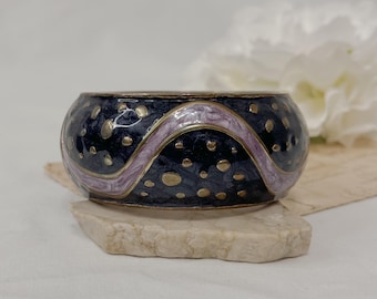 Può includere: Un braccialetto nero e oro con un designo in smalto viola e bianco. Il braccialetto ha un motivo ondulato ed è decorato con piccoli punti dorati.