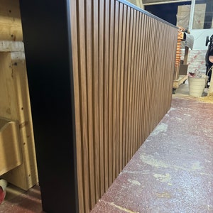 May include: A large, rectangular wall panel made of vertical wooden slats with a black painted border. The wood is a rich brown colour and the slats are evenly spaced.