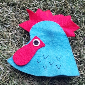 May include: A felt rooster puppet with a red comb and wattle, blue body, and black eyes. The puppet is lying on a bed of green grass.