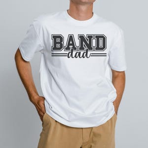 May include: White t-shirt with the text "BAND dad" in a black, collegiate-style font. The word "BAND" is in a bold, block letter style, and "dad" is in a script font. The shirt is a crew neck style.