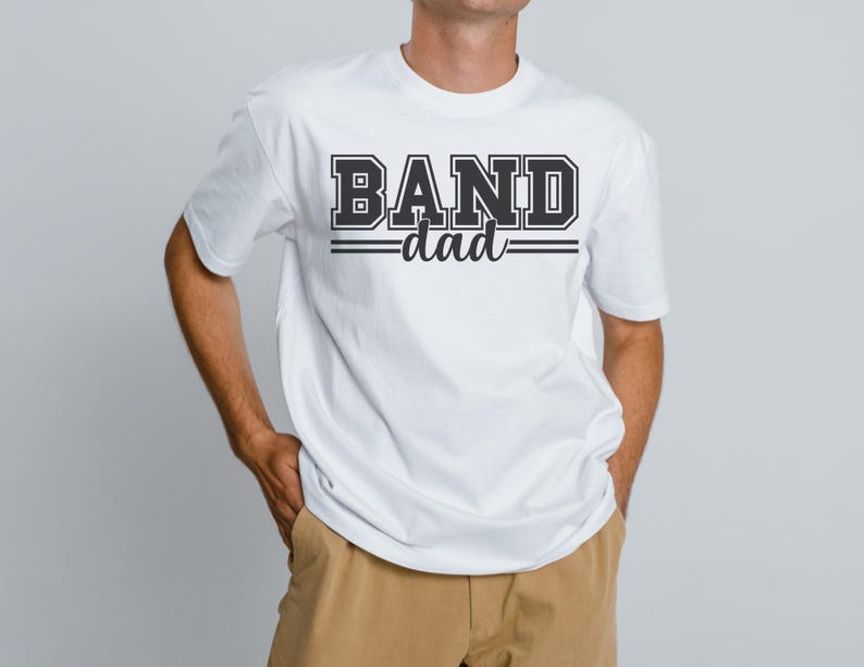 May include: White t-shirt with the text "BAND dad" in a black, collegiate-style font. The word "BAND" is in a bold, block letter style, and "dad" is in a script font. The shirt is a crew neck style.