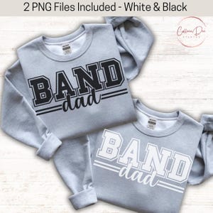May include: Two light gray sweatshirts with the text "BAND dad" in a collegiate font. One sweatshirt has black lettering, and the other has white lettering. The sweatshirts are displayed on a light-colored surface.
