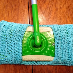 Puede incluir: Una funda de fregona de ganchillo azul claro. La funda es rectangular y se adapta a un cabezal de fregona Swiffer verde. El punto de ganchillo crea un patrón texturizado. El mango de la fregona es verde y plateado.