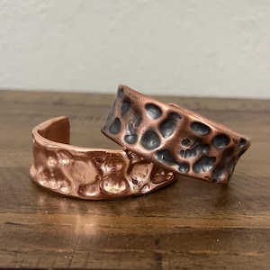 May include: Two copper cuff bracelets with a hammered texture. One bracelet is lying flat, while the other is slightly angled, showcasing the detailed surface. The bracelets have an open design and a warm, metallic finish.