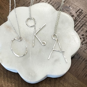 May include: Three silver-colored initial necklaces, spelling out the letters "C", "K", and "A". Each letter is crafted from a single wire and hangs from a delicate chain. The necklaces are displayed on a white, flower-shaped surface.