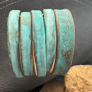 May include: A cuff bracelet with a weathered, turquoise patina. The bracelet is made of five curved, vertical segments, each with a copper-colored edge. The metal has a textured appearance, suggesting age and wear. The bracelet is a statement piece.