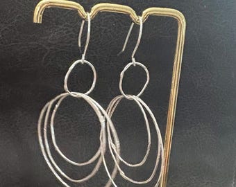 Puede incluir: Pendientes colgantes plateados con un diseño ovalado en capas. Cada pendiente presenta una parte superior pequeña y redonda y tres óvalos texturizados más grandes. Los pendientes se exhiben en un expositor de joyería dorado sobre un fondo oscuro.