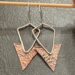 May include: Geometric earrings with a hammered copper triangle and silver-tone metal frame. The earrings are suspended from a silver wire hook. The copper has a textured surface and a reddish-brown color.
