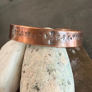 May include: A copper cuff bracelet featuring stamped text that reads N32.8 35 E and W 96.766, separated by a small compass rose symbol. The metal has a warm, reddish-brown metallic finish with a smooth, slightly curved band design.