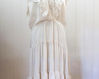 Puede incluir: Un vestido vintage color crema con escote en V y falda escalonada. El vestido presenta un delicado cuello y ribete de encaje, y se exhibe en un maniquí sobre un fondo blanco. El vestido tiene un diseño fluido y escalonado.