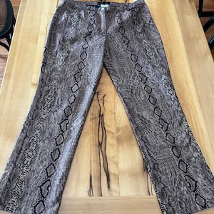 May include: Full-length trousers featuring a brown and black snakeskin pattern. The trousers have a straight-leg cut, a central button and zip fly closure, and a waistband with belt loops. The fabric displays a textured, reptilian-inspired print.